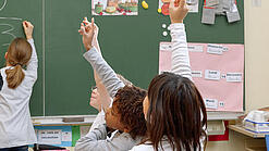 Eine Lehrerin steht vor einer Tafel, Schülerinnen melden sich.