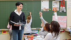 Eine Lehrerin steht vor einer Tafel, Schülerinnen melden sich.