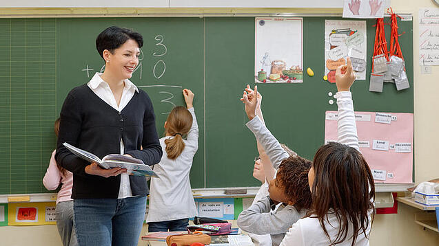 Eine Lehrerin steht vor einer Tafel, Schülerinnen melden sich.