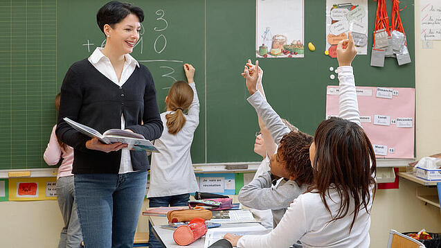 Eine Lehrerin steht vor einer Tafel, Schülerinnen melden sich.