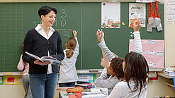Eine Lehrerin steht vor einer Tafel, Schülerinnen melden sich.