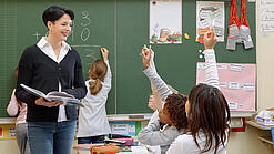 Eine Lehrerin steht vor einer Tafel, Schülerinnen melden sich.