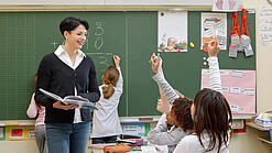 Eine Lehrerin steht vor einer Tafel, Schülerinnen melden sich.