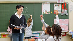 Eine Lehrerin steht vor einer Tafel, Schülerinnen melden sich.