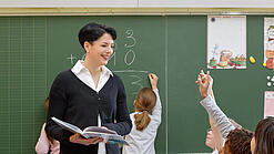 Eine Lehrerin steht vor einer Tafel, Schülerinnen melden sich.