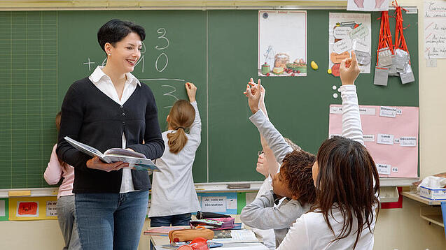 Eine Lehrerin steht vor einer Tafel, Schülerinnen melden sich.
