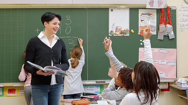 Eine Lehrerin steht vor einer Tafel, Schülerinnen melden sich.