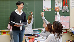 Eine Lehrerin steht vor einer Tafel, Schülerinnen melden sich.