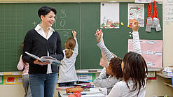 Eine Lehrerin steht vor einer Tafel, Schülerinnen melden sich.