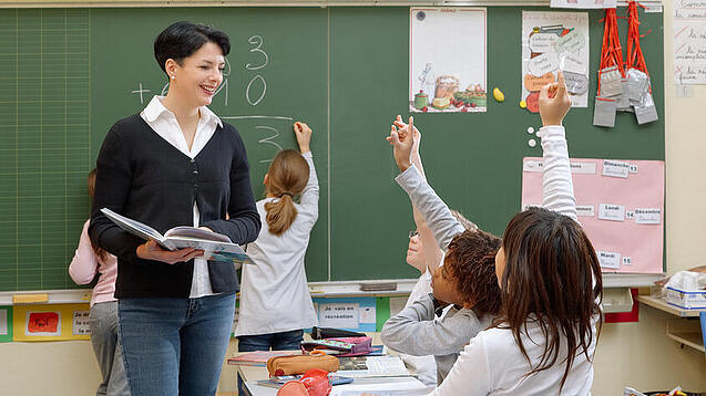 Eine Lehrerin steht vor einer Tafel, Schülerinnen melden sich.