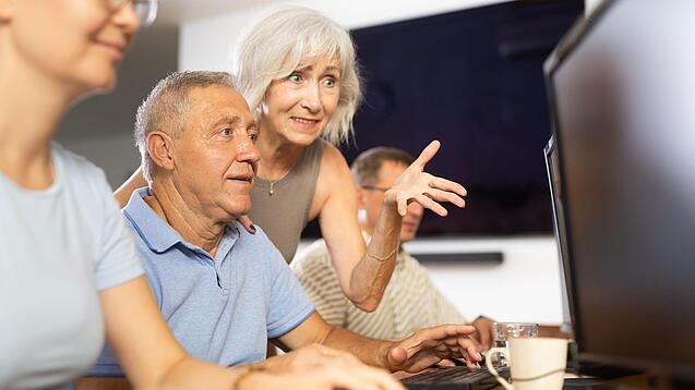 Ein älterer Mann sitzt vor einem PC und eine ältere Frau erklärt ihm etwas.