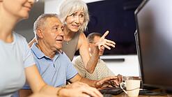 Ein älterer Mann sitzt vor einem PC und eine ältere Frau erklärt ihm etwas.