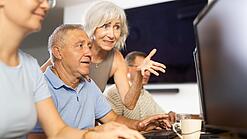Ein älterer Mann sitzt vor einem PC und eine ältere Frau erklärt ihm etwas.