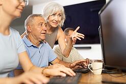 Ein älterer Mann sitzt vor einem PC und eine ältere Frau erklärt ihm etwas.
