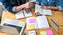 Zwei Personen sitzen an einem Tisch, auf dem ein Laptop,Bücher und Notzihefte liegen.