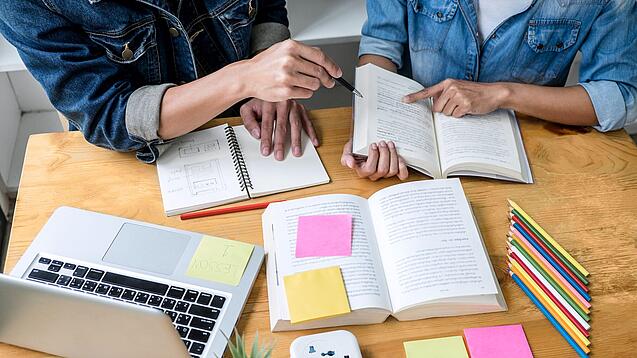 Zwei Personen sitzen an einem Tisch, auf dem ein Laptop,Bücher und Notzihefte liegen.
