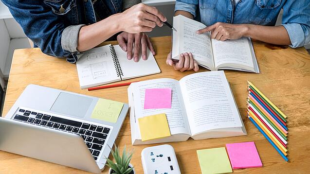 Zwei Personen sitzen an einem Tisch, auf dem ein Laptop,Bücher und Notzihefte liegen.