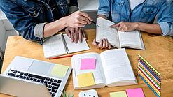 Zwei Personen sitzen an einem Tisch, auf dem ein Laptop,Bücher und Notzihefte liegen.