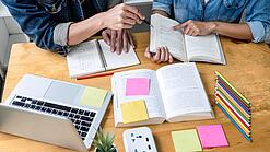 Zwei Personen sitzen an einem Tisch, auf dem ein Laptop,Bücher und Notzihefte liegen.