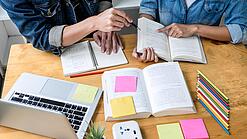 Zwei Personen sitzen an einem Tisch, auf dem ein Laptop,Bücher und Notzihefte liegen.