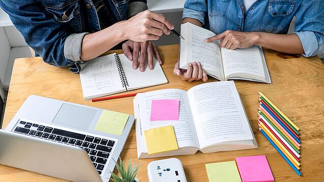 Zwei Personen sitzen an einem Tisch, auf dem ein Laptop,Bücher und Notzihefte liegen.