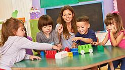 Kinder sitzen mit einer Erzieherin am Tisch und spielen.
