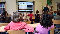Kinder sitzen in einem Klassenraum und schauen auf ein digitales Whiteboard.