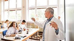 Chemielehrer erklärt Jugendlichen etwas an der Tafel.