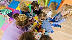 Eine Erzieherin sitzt mit Kindergartenkindern in einem Kreis und schaut auf Buchstaben. 