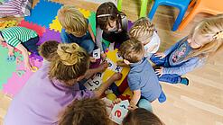 Eine Erzieherin sitzt mit Kindergartenkindern in einem Kreis und schaut auf Buchstaben. 