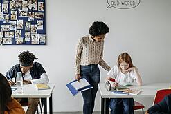 In einem Klassenzimmer steht eine Lehrerin bei einer Schülerin am Tisch und erklärt ihr etwas. Eine Junge arbeitet konzentriert an einem Tisch daneben.