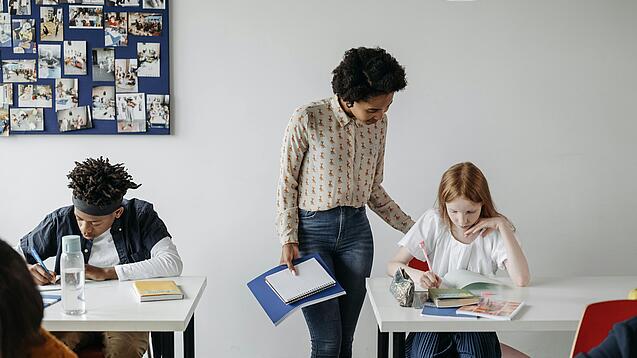 In einem Klassenzimmer steht eine Lehrerin bei einer Schülerin am Tisch und erklärt ihr etwas. Eine Junge arbeitet konzentriert an einem Tisch daneben.
