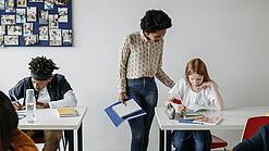 In einem Klassenzimmer steht eine Lehrerin bei einer Schülerin am Tisch und erklärt ihr etwas. Eine Junge arbeitet konzentriert an einem Tisch daneben.