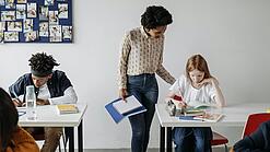 In einem Klassenzimmer steht eine Lehrerin bei einer Schülerin am Tisch und erklärt ihr etwas. Eine Junge arbeitet konzentriert an einem Tisch daneben.