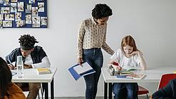 In einem Klassenzimmer steht eine Lehrerin bei einer Schülerin am Tisch und erklärt ihr etwas. Eine Junge arbeitet konzentriert an einem Tisch daneben.