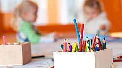 Stifte stehen auf dem Tisch einer Kita, im Hintergrund zwei Kinder.