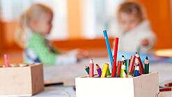 Stifte stehen auf dem Tisch einer Kita, im Hintergrund zwei Kinder.