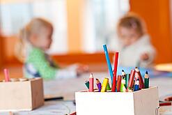 Stifte stehen auf dem Tisch einer Kita, im Hintergrund zwei Kinder.