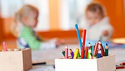 Stifte stehen auf dem Tisch einer Kita, im Hintergrund zwei Kinder.