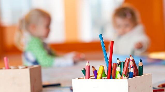 Stifte stehen auf dem Tisch einer Kita, im Hintergrund zwei Kinder.