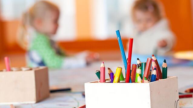 Stifte stehen auf dem Tisch einer Kita, im Hintergrund zwei Kinder.