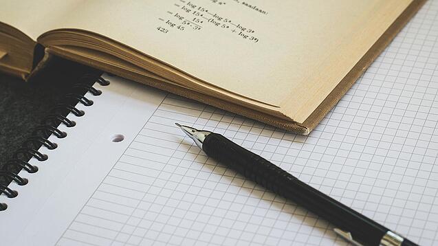 Ein Buch liegt aufgeschlagen auf einem Notizblock, auf dem auch ein Stift liegt.