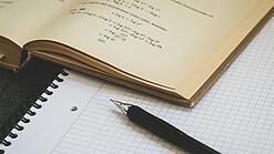 Ein Buch liegt aufgeschlagen auf einem Notizblock, auf dem auch ein Stift liegt.