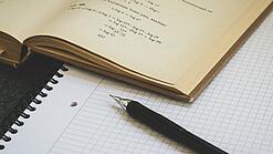 Ein Buch liegt aufgeschlagen auf einem Notizblock, auf dem auch ein Stift liegt.