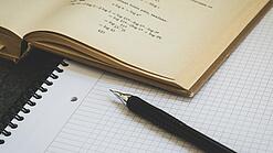 Ein Buch liegt aufgeschlagen auf einem Notizblock, auf dem auch ein Stift liegt.