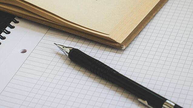 Ein Buch liegt aufgeschlagen auf einem Notizblock, auf dem auch ein Stift liegt.