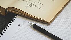 Ein Buch liegt aufgeschlagen auf einem Notizblock, auf dem auch ein Stift liegt.