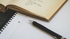 Ein Buch liegt aufgeschlagen auf einem Notizblock, auf dem auch ein Stift liegt.