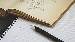 Ein Buch liegt aufgeschlagen auf einem Notizblock, auf dem auch ein Stift liegt.