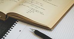 Ein Buch liegt aufgeschlagen auf einem Notizblock, auf dem auch ein Stift liegt.